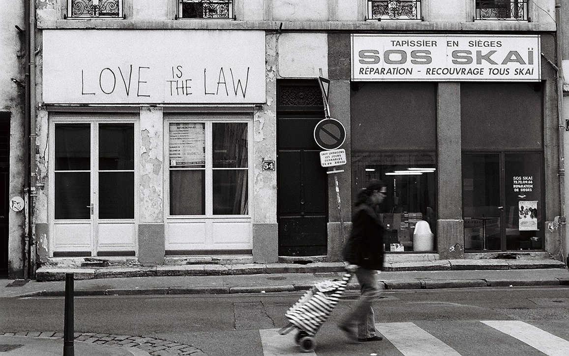 Photographie de rue montrant une passante traversant devant un magasin "SOS Skai" à côté d'un graffiti "Love is the law", à Lyon